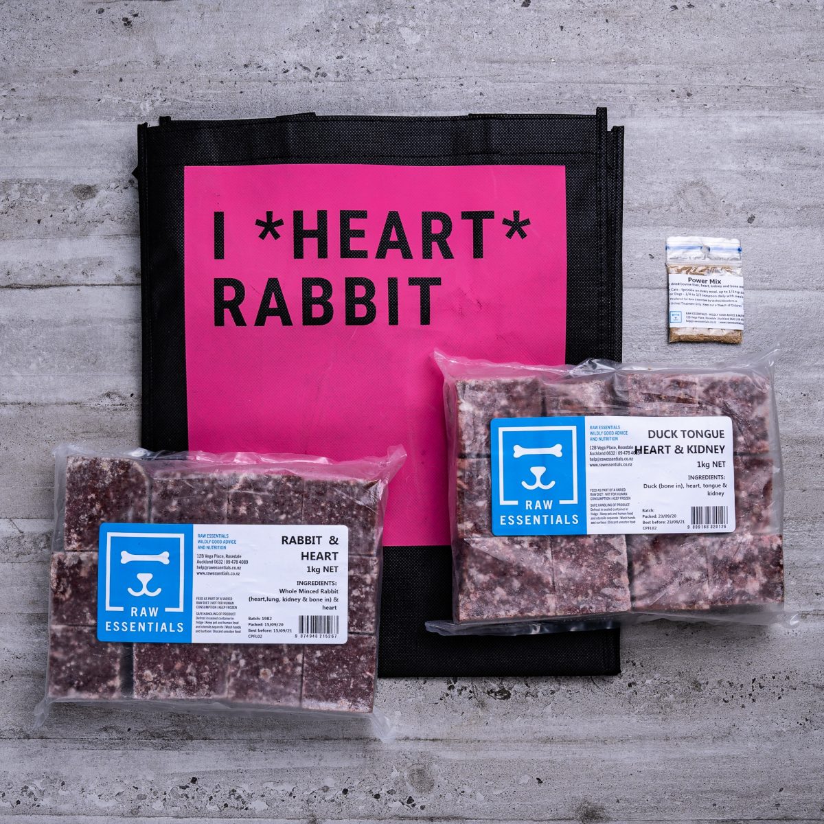 Assortment Of Raw Essential Products For Cats Including, Pink And Black Tote Bag With "I *Heart* Rabbit" Written,  Sample Bag Of Power Mix, 1KG Plastic Pack Of Cubed Rabbit Heart Meat Mix, 1KG Plastic Pack Of Cubed Duck Tongue, Heart And Kidney Meat Mix