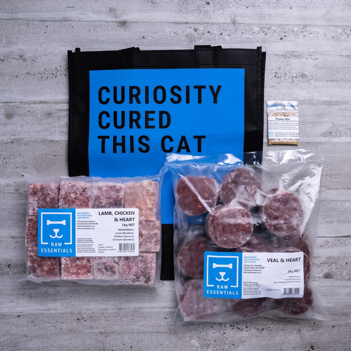 Assortment Of Raw Essential Products For Cats Including, Blue And Black Tote Bag With "Curiosity Cured This Cat" Written,  Sample Bag Of Power Mix, 1KG Plastic Pack Of Veal And Heart Mix , 1KG Plastic Pack Of Cubed Lamb, Chicken, Heart Mix.