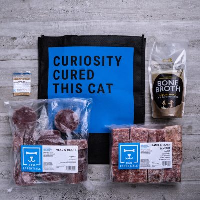 Assortment Of Raw Essential Products For Kittens Including, Blue And Black Tote Bag With "Curiosity Cured This Cat" Written, Bone Broth,  Sample Bag Of Power Mix, 1KG Plastic Pack Of Veal And Heart Mix , 1KG Plastic Pack Of Cubed Lamb, Chicken, Heart Mix.